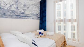 Una cama bien hecha con almohadas y una bandeja con una taza y vasos, un ventanal con cortinas traslúcidas y un mural en la pared con barcos a vela.