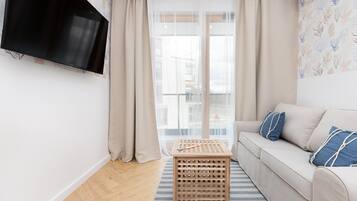 Una sala de estar con un sofá, un televisor montado en la pared, una mesa de madera y una ventana con cortinas de gasa.