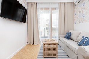 Una sala de estar con un sofá, un televisor montado en la pared, una mesa de madera y una ventana con cortinas de gasa.