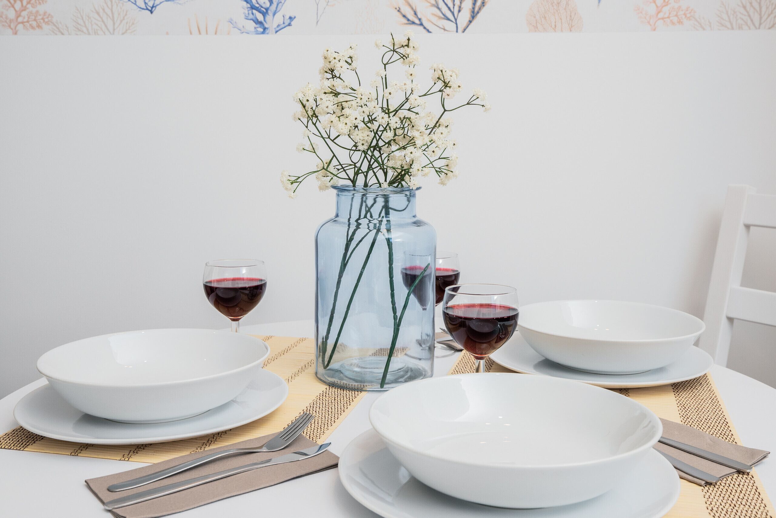Een eettafel met witte borden, kommen en glazen rode wijn, een heldere glazen vaas met witte bloemen, en een geweven placemat.