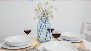 Una mesa de comedor con platos blancos, tazones, copas de vino tinto y un florero de vidrio transparente con flores blancas, además de un mantel tejido.