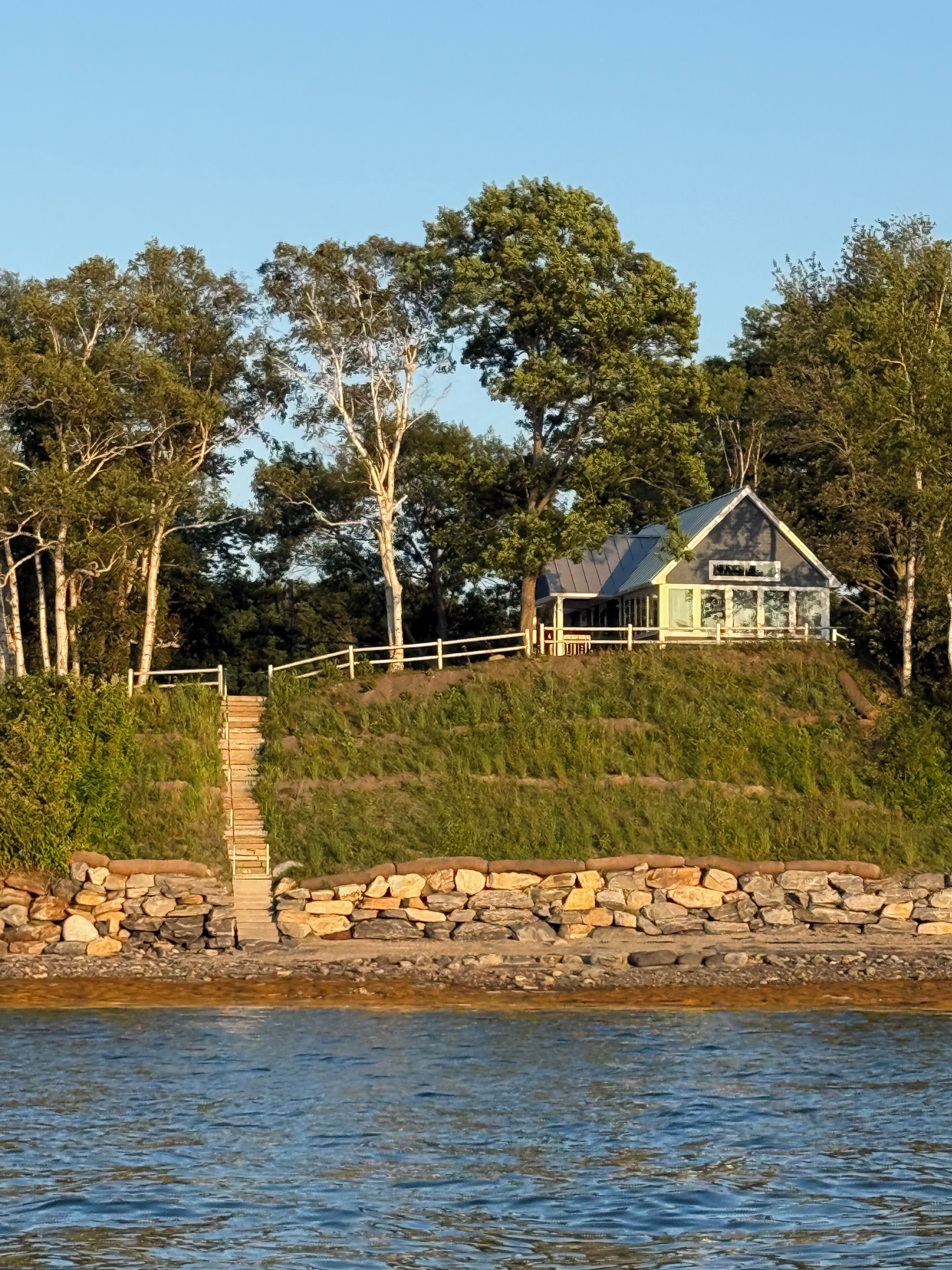 Une maison avec un chemin de pierre menant à une terrasse en bois, entourée d’arbres et donnant sur une étendue d’eau.