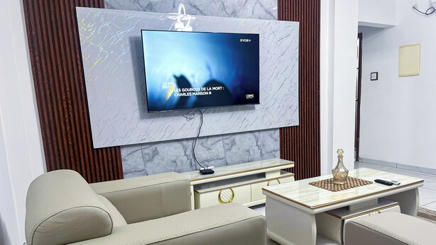 Una sala de estar moderna con un televisor de pantalla plana montado en una pared de mármol, un sofá color beige, una mesa de centro con un objeto decorativo y una pequeña mesita auxiliar.