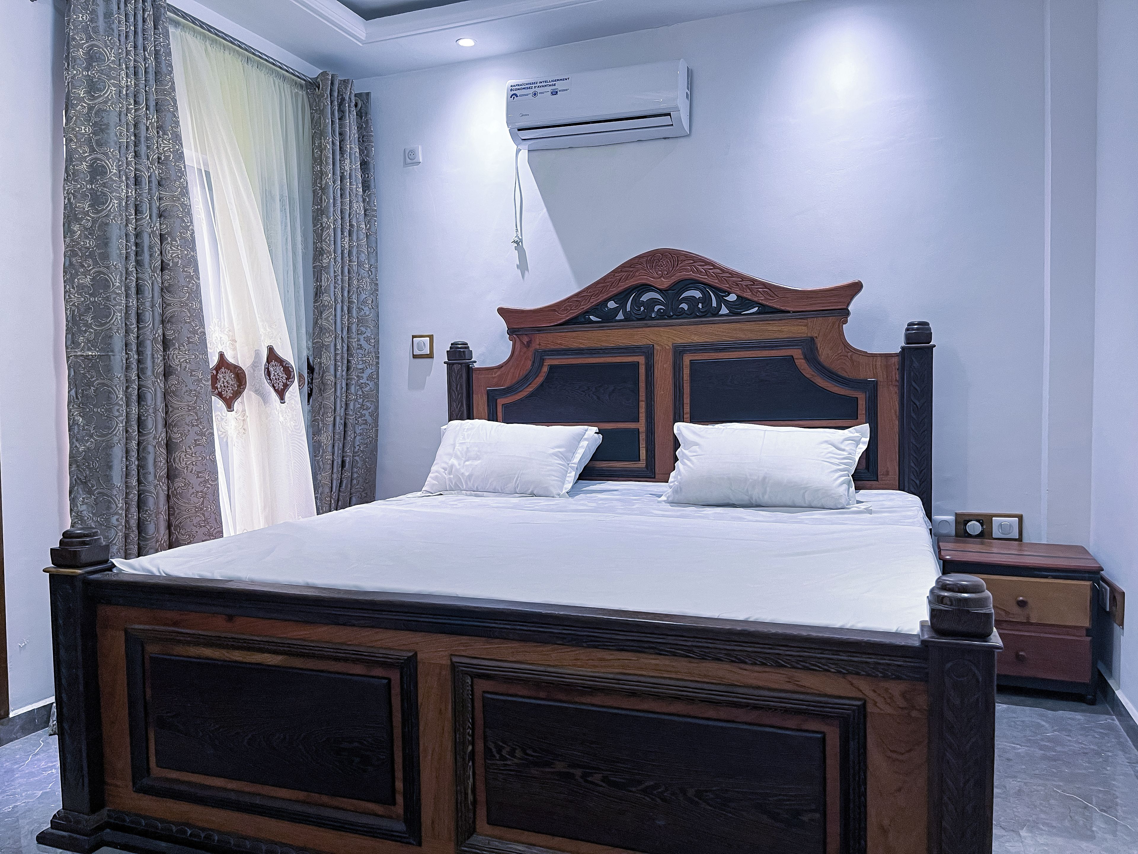 A bedroom with a wooden bed, white bedding, a nightstand, an air conditioning unit, and patterned curtains.