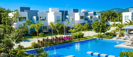 A modern hotel with a swimming pool, surrounded by lush greenery and a clear blue sky.