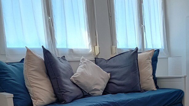 A white sofa with blue and beige cushions, positioned in front of a window with sheer curtains.