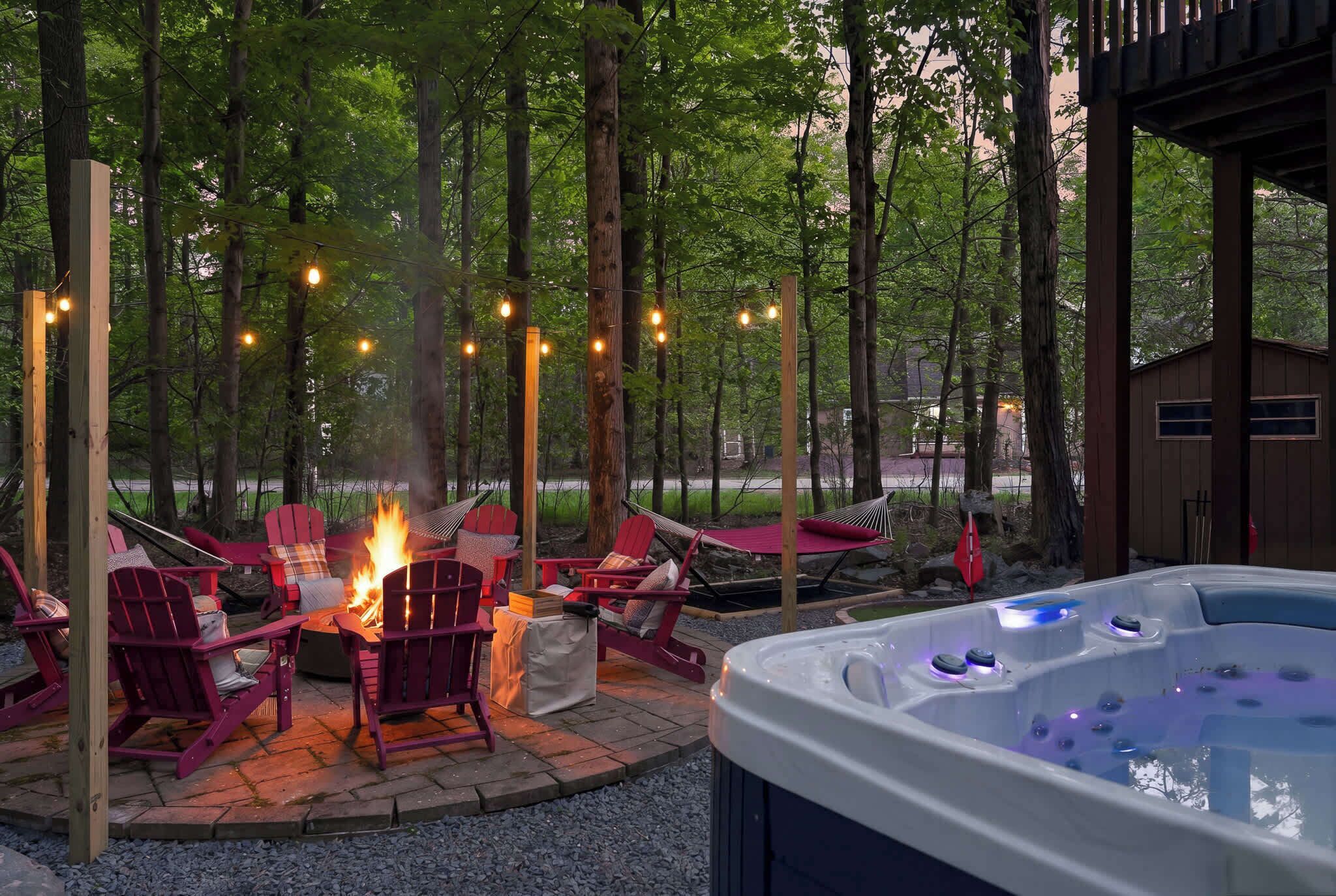 Ein Whirlpool mit Feuerstelle und roten Adirondack-Stühlen in einer bewaldeten Außenanlage.