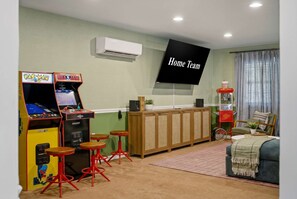 Ein Zimmer mit Spielautomaten, einem Fernsehbildschirm, der 'Home Team' zeigt, und einer Sitzecke mit einer Couch und einem Couchtisch.