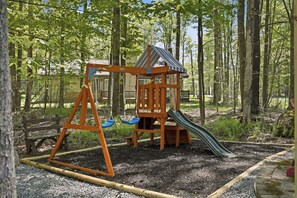 Ein Spielplatz mit Schaukel, Rutsche und Klettergerüst, umgeben von Bäumen.