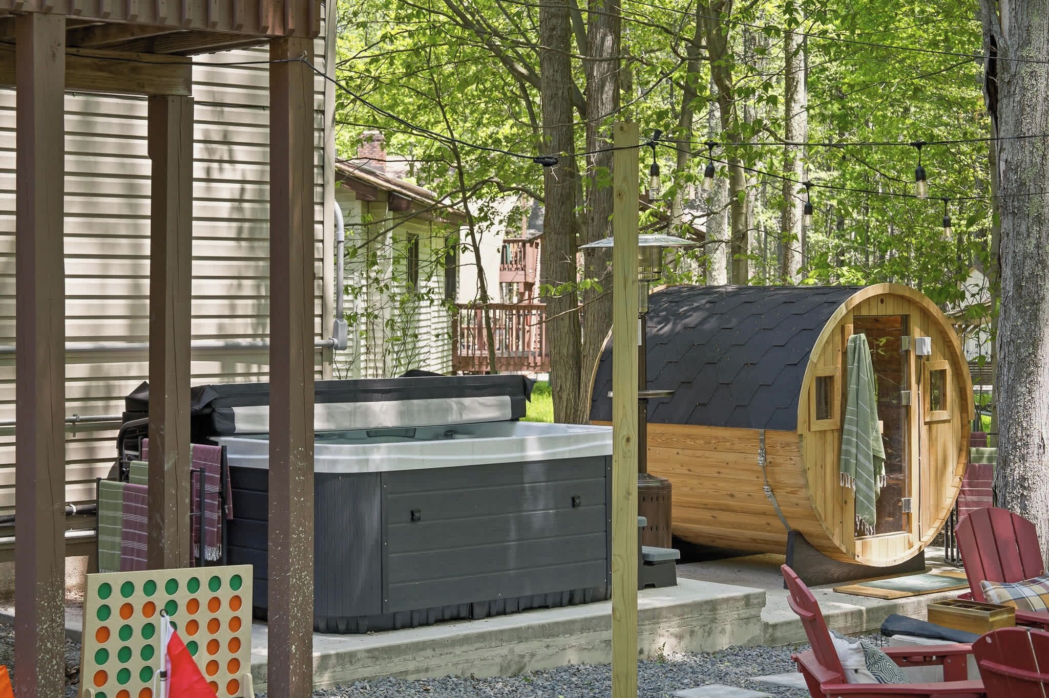 A hot tub and a wooden cabin in a wooded area with a deck and chairs.