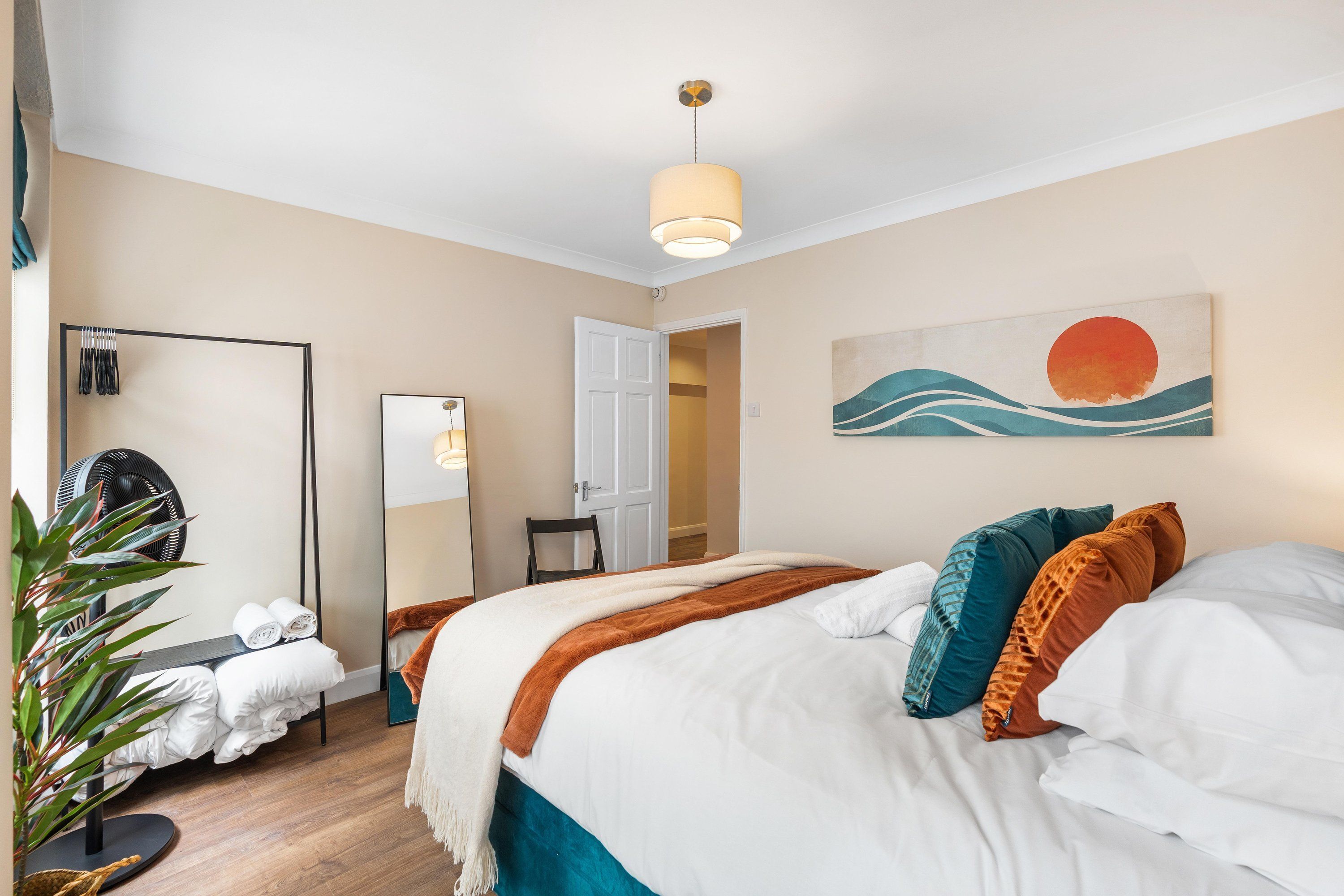 A hotel room with a bed, a mirror, a fan, a plant, and a painting on the wall.