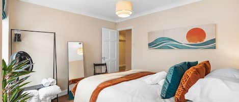 A hotel room with a bed, a mirror, a fan, a plant, and a painting on the wall.