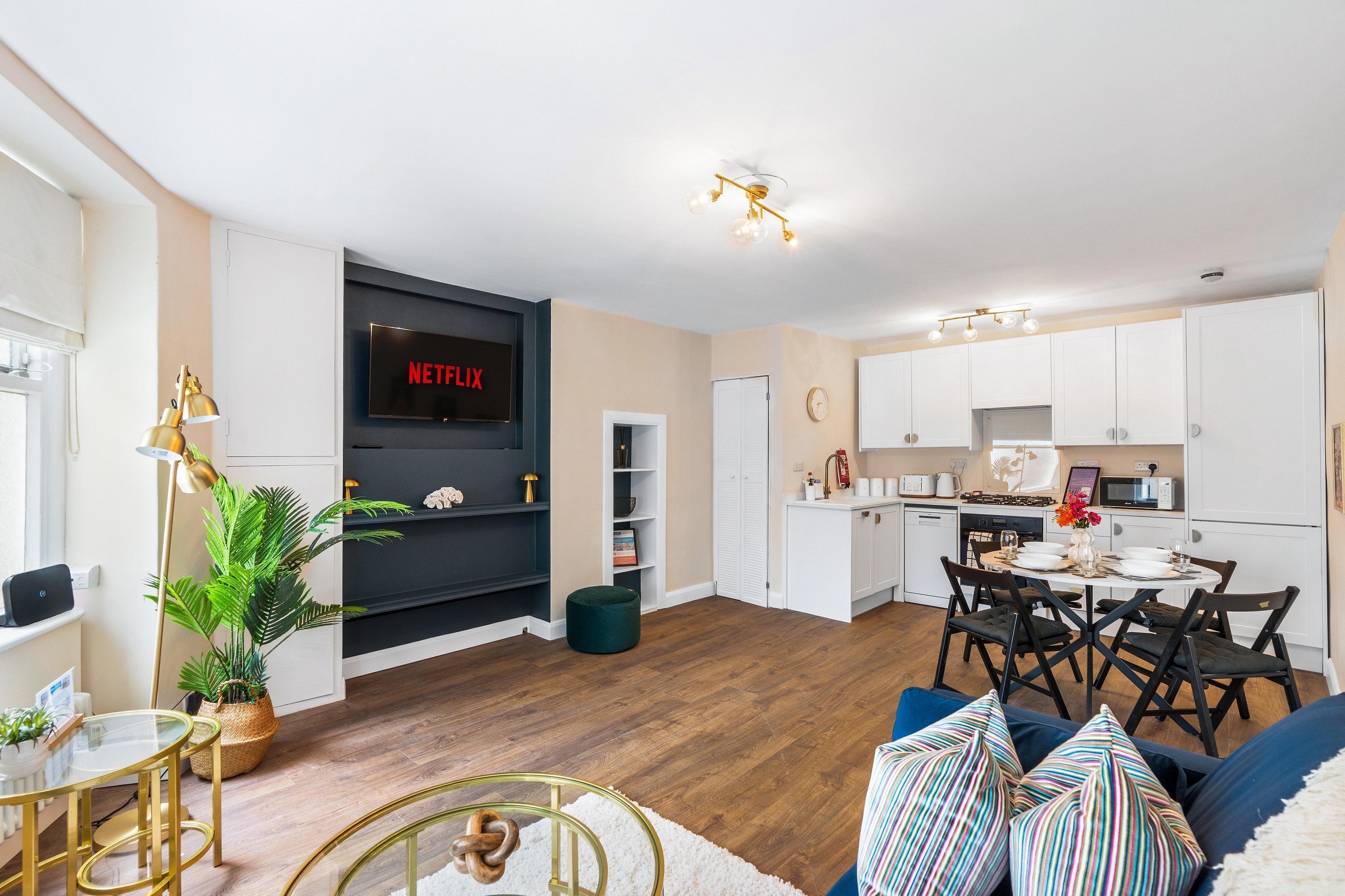 A modern living room with a flat-screen TV, a dining area with a table and chairs, and a kitchen with white cabinets.