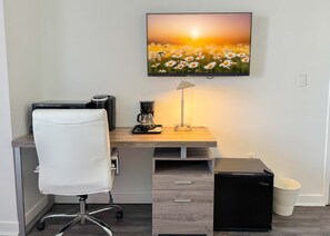 Ein modernes Büro mit Schreibtisch, Stuhl, Kaffeemaschine, Lampe und einem Fernseher, der eine Blumenwiese zeigt.