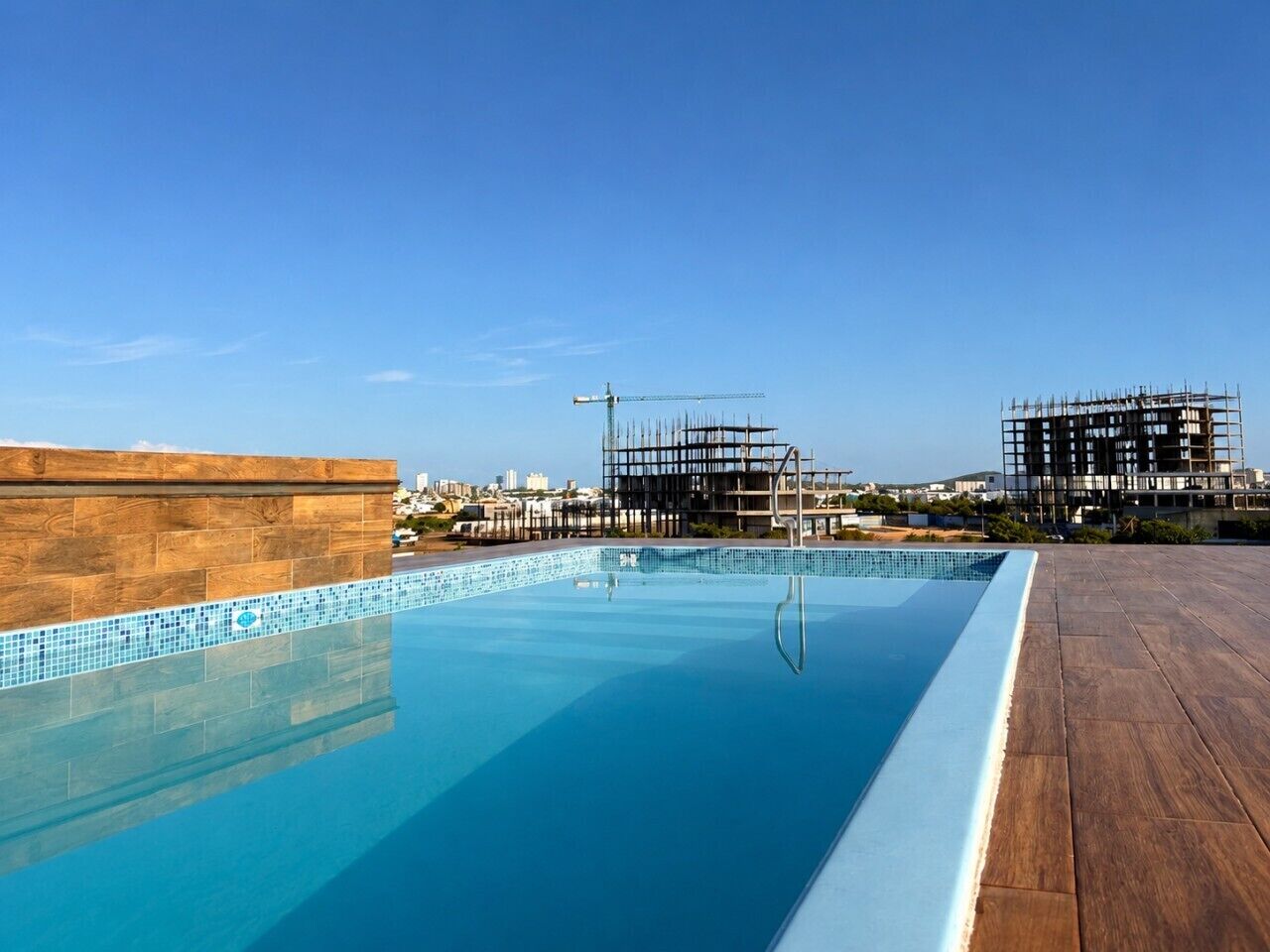 A rooftop swimming pool with a wooden deck, a clear blue sky, and a construction site in the background.