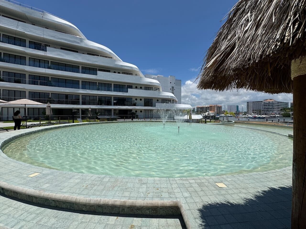Ein Swimmingpool mit Springbrunnen, umgeben von einer gefliesten Terrasse, mit einem modernen, mehrstöckigen Gebäude im Hintergrund.