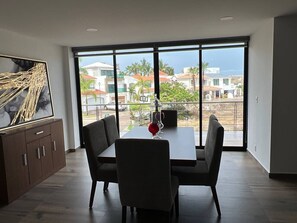 Ein Essbereich mit Tisch und Stühlen, ein großes Fenster mit Blick auf Gebäude und Palmen, sowie eine Kommode mit Schubladen.