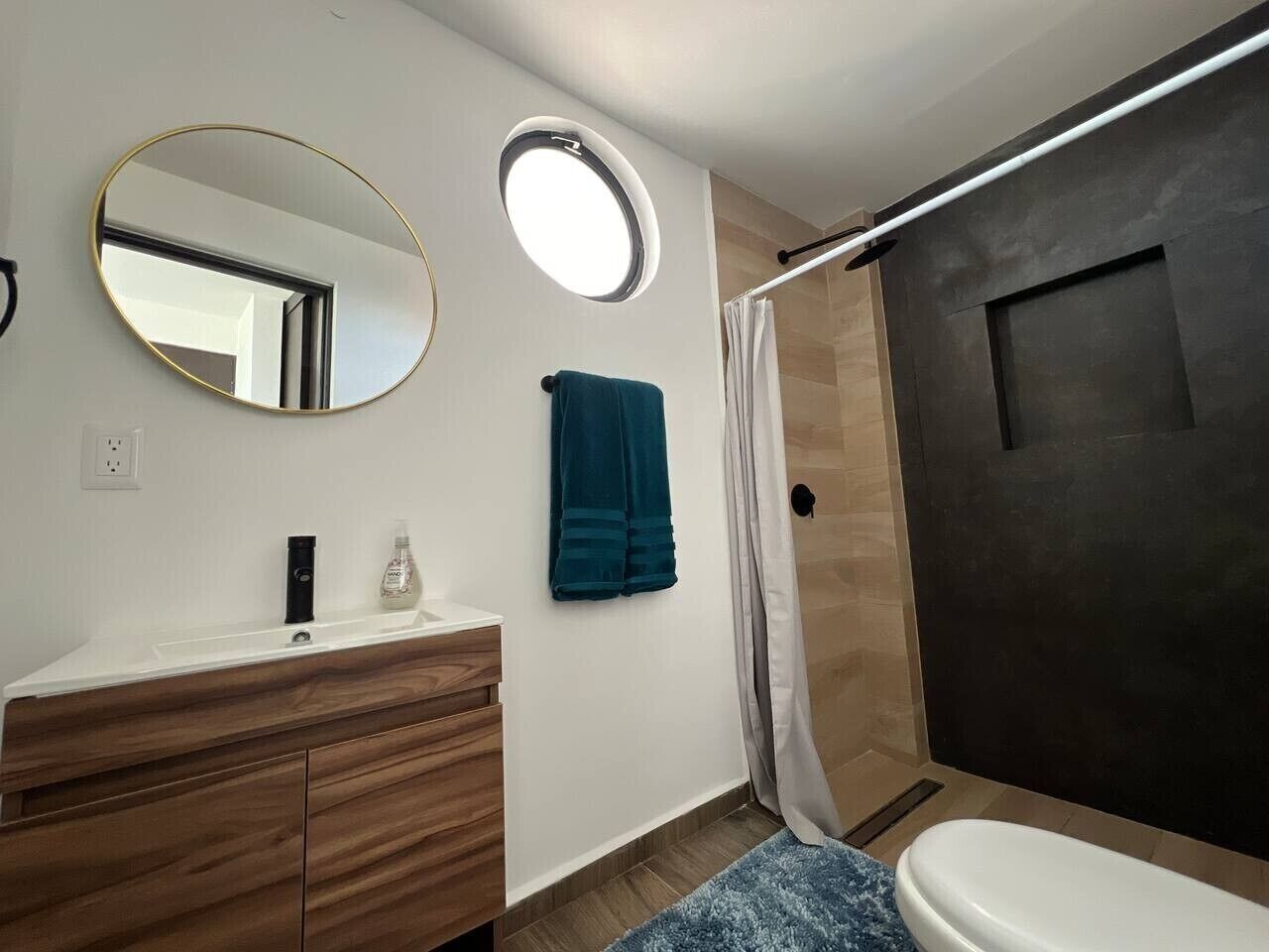A bathroom with a round mirror, a sink with a wooden vanity, a towel, and a shower area.