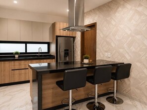 Eine moderne Küche mit einer zentralen Kochinsel, Barhocker, einem Edelstahlkühlschrank und einer Küchenrückwand mit Fischgrätenmuster.