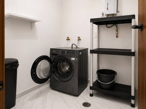 A compact laundry room with a front-loading washing machine, a black trash bin, and a metal shelf unit with a black bucket. - Ubicación ideal | depto. nuevo con 2 recámaras (Mazatlán)