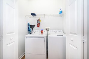 A laundry room with two white front-loading washing machines, a blue detergent bottle, a fire extinguisher, and a blue water jug on a shelf. - 4-Bedroom Pool Home Near Disney | 2 Master Suites | Sleeps 10 (Clermont)