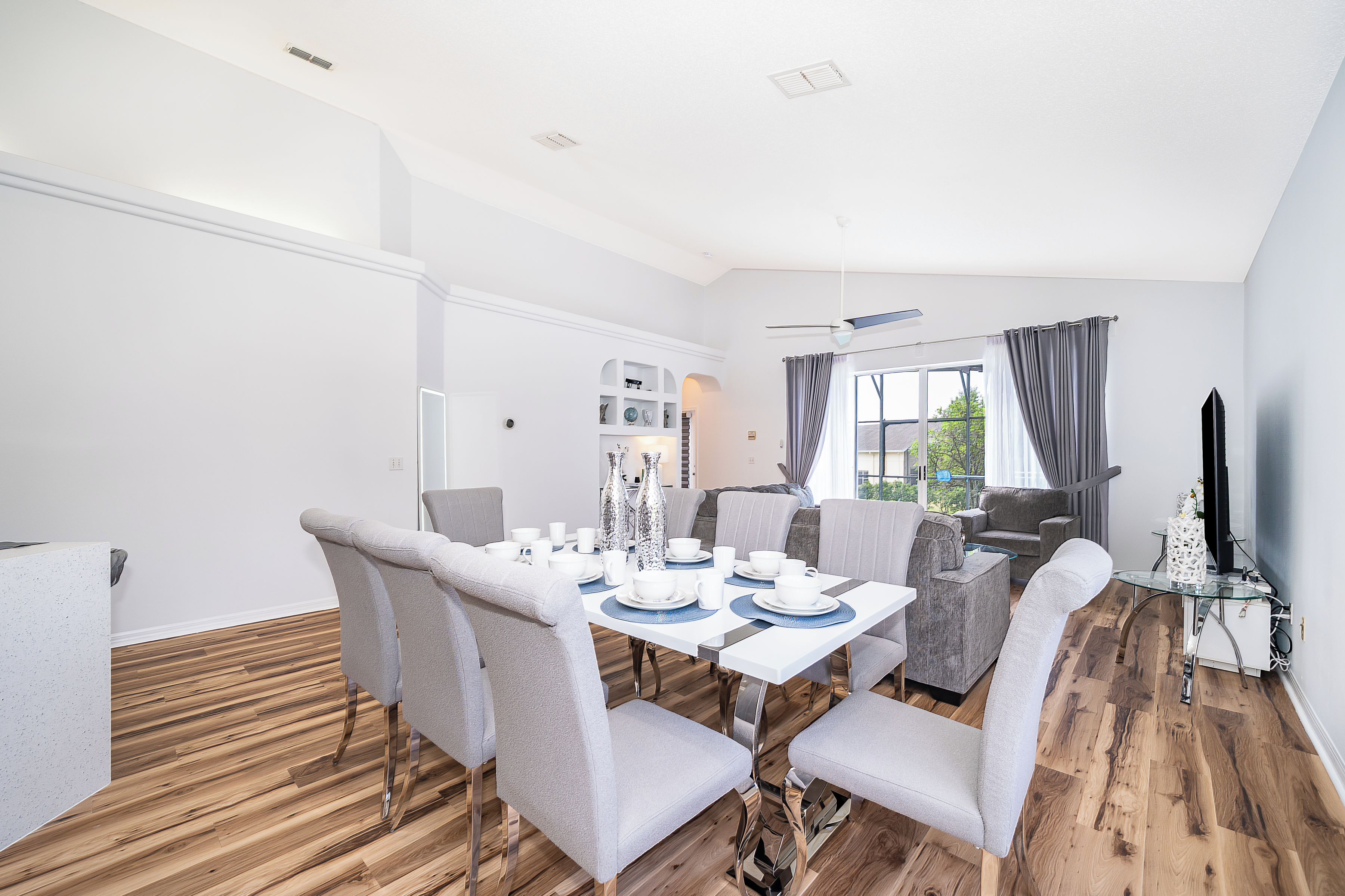 A modern dining area with a white table set for six, grey upholstered chairs, and a wooden floor.