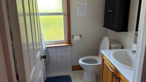 Une petite salle de bain avec des toilettes, un lavabo et un meuble. Il y a une fenêtre donnant sur un champ verdoyant. Un panneau sur le mur demande de ne pas jeter de serviettes en papier ni de protections hygiéniques dans les toilettes.
