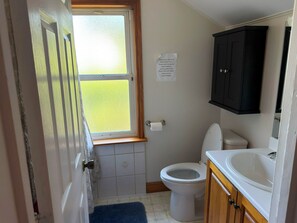 Un baño pequeño con inodoro, lavamanos y armario. Hay una ventana con vistas a un campo verde. Un cartel en la pared solicita no desechar toallas de papel ni compresas en el inodoro.
