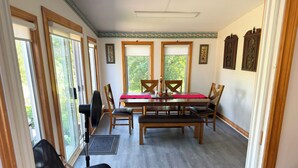 Un espace repas comprenant une table, des chaises et un banc. Il y a des fenêtres avec des stores, un ventilateur et une porte avec un rideau.