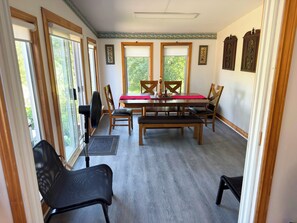 Un comedor con una mesa, sillas y un banco. Hay ventanas con persianas, un ventilador y una puerta con cortina.