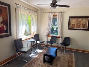 Una habitación con ventilador de techo, dos ventanas con cortinas, un cuadro y una pequeña mesa con sillas.