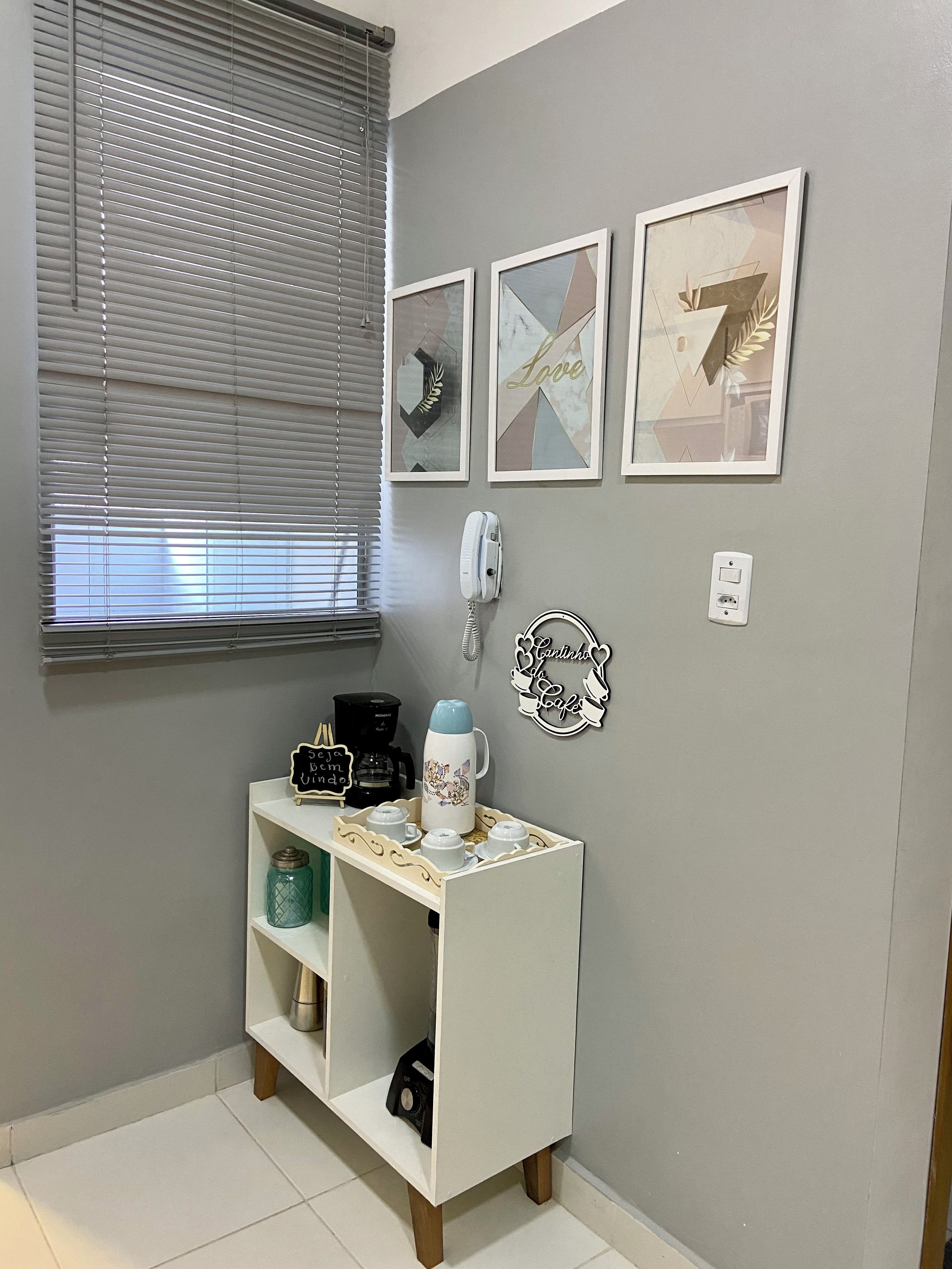 A room with a white console table, a coffee maker, a kettle, and a shelf with a jar. There are three framed artworks on the wall and a wall-mounted phone.