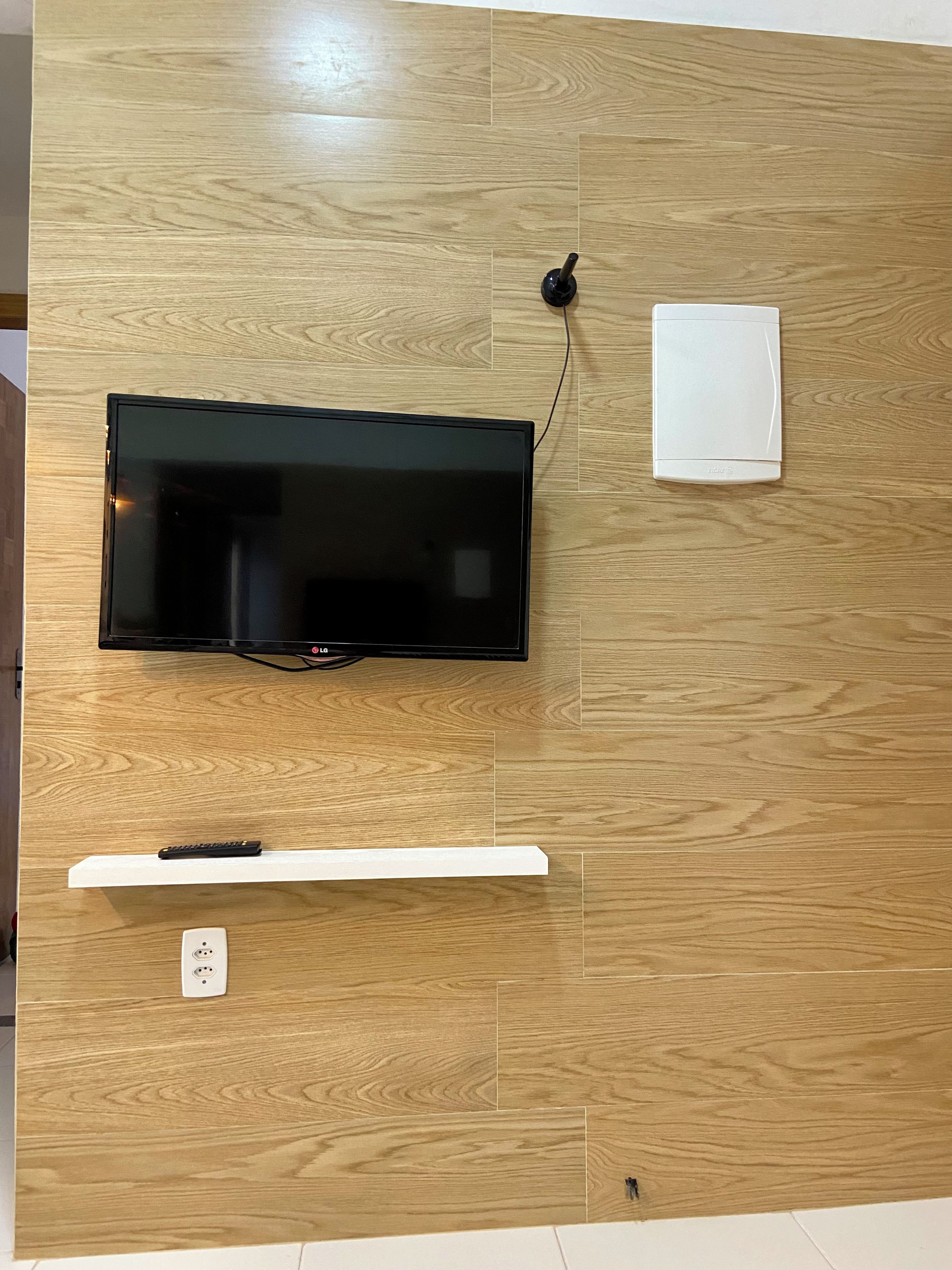 Televisor de pantalla plana montado en una pared de madera, con un cable, un enchufe y un estante blanco debajo.
