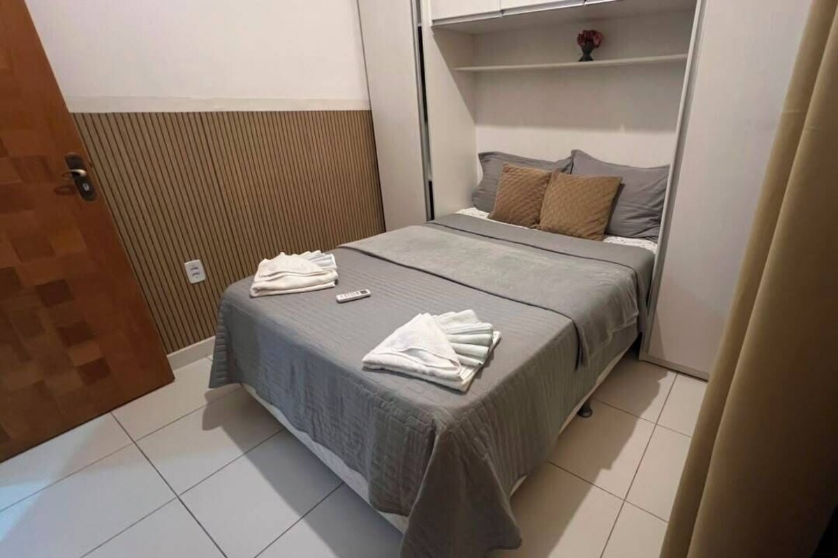Quarto de hotel com cama, mesa de cabeceira, prateleira na parede e porta.