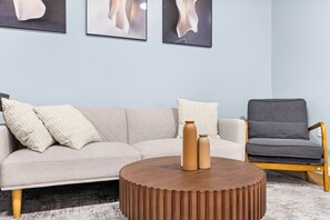 Ein Wohnzimmer mit einer Couch, einem runden Holztisch, zwei Vasen, einem grauen Sessel und abstrakter Wandkunst.