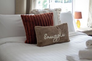 Cama bem arrumada com lençóis brancos e duas almofadas decorativas, uma delas com a palavra 'Snuggle'.