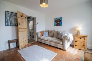 A cozy living room with a sofa, a wooden cabinet, and a door leading to a staircase. - Ruby Cottage | Garden & Wood Burner Near Stamford (Easton on the Hill)