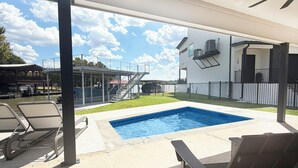 Zona de piscina exterior con un porche cubierto, sillas de descanso y vistas a un edificio moderno con fachada blanca.