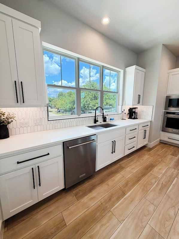 A modern kitchen with white cabinetry, a stainless steel dishwasher, and a large window with a view of greenery.