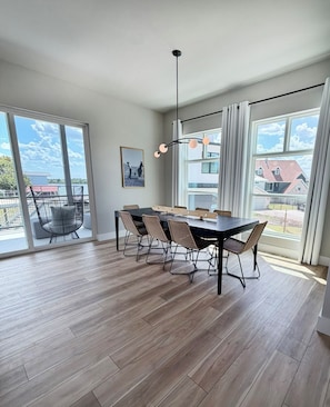 A spacious dining area with a large table and chairs, large windows with curtains, and a view of a residential area.