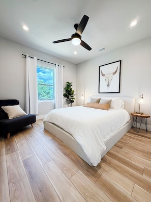 A modern bedroom with a bed, a ceiling fan, a window with curtains, a plant, and a framed artwork on the wall.