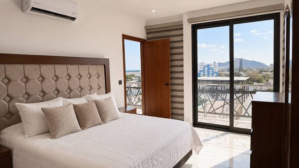 A bedroom with a large bed, a wooden headboard, a city view through a sliding door, and a built-in air conditioning unit. - Dpto. nuevo a unos pasos de malecón y zona dorada (Mazatlán)