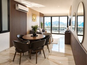 A modern dining area with a round table and chairs, a potted plant, and a city view through large windows. - Dpto. nuevo a unos pasos de malecón y zona dorada (Mazatlán)