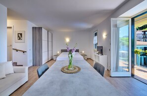 A modern hotel room with a long dining table, a sofa, a TV, and a balcony with plants. - Domus Lario's Lake – stunning view, beach, swimming pool, and private parking (Lierna)