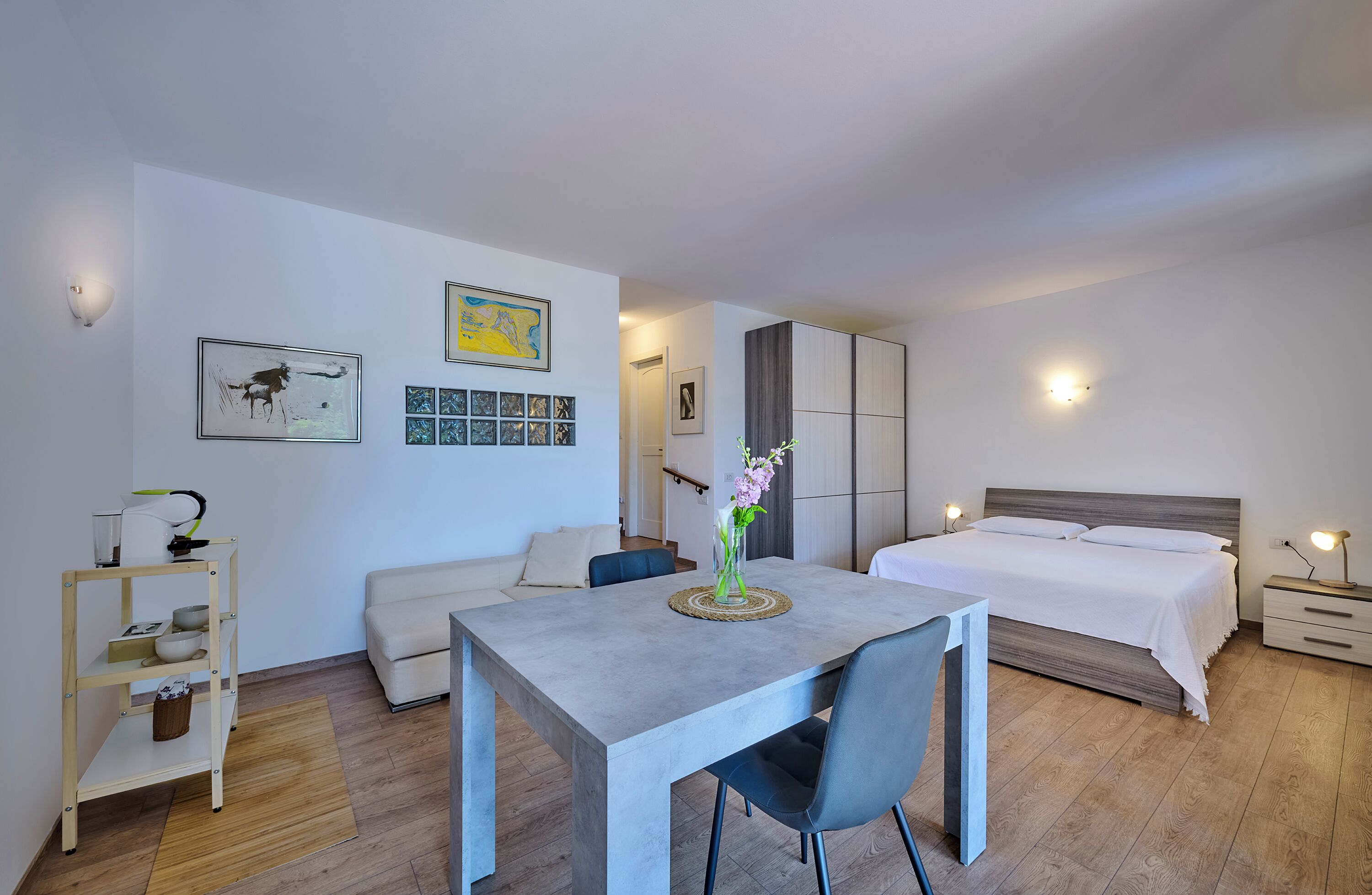 A modern bedroom with a bed, bedside tables, a desk, and a chair. There are wall-mounted lights, a framed picture, and a vase with flowers on the desk.