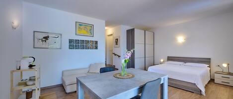 A modern bedroom with a bed, bedside tables, a desk, and a chair. There are wall-mounted lights, a framed picture, and a vase with flowers on the desk.
