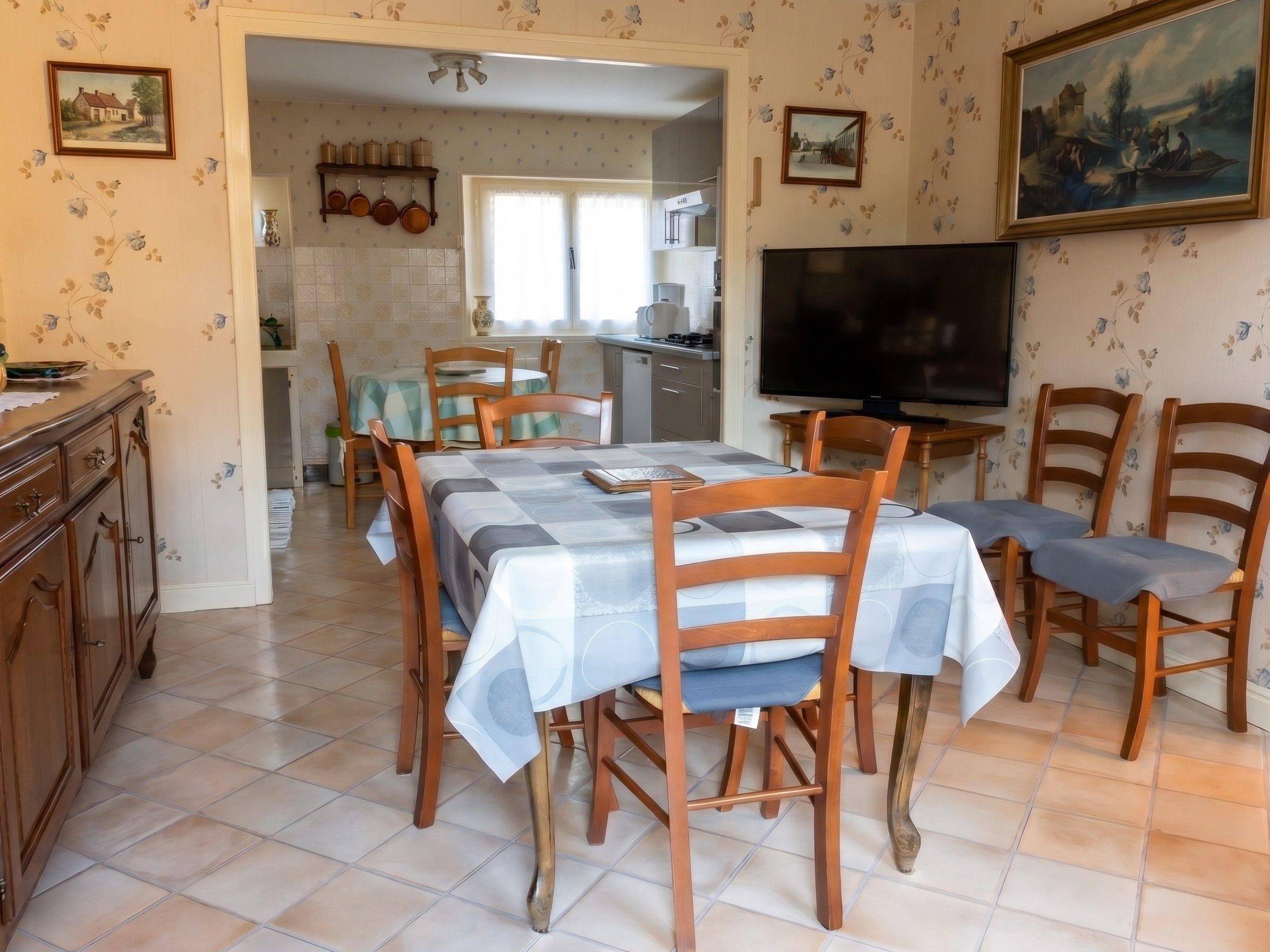 A dining area with a table set for four, surrounded by wooden chairs with cushioned seats. A television is mounted on the wall to the right.