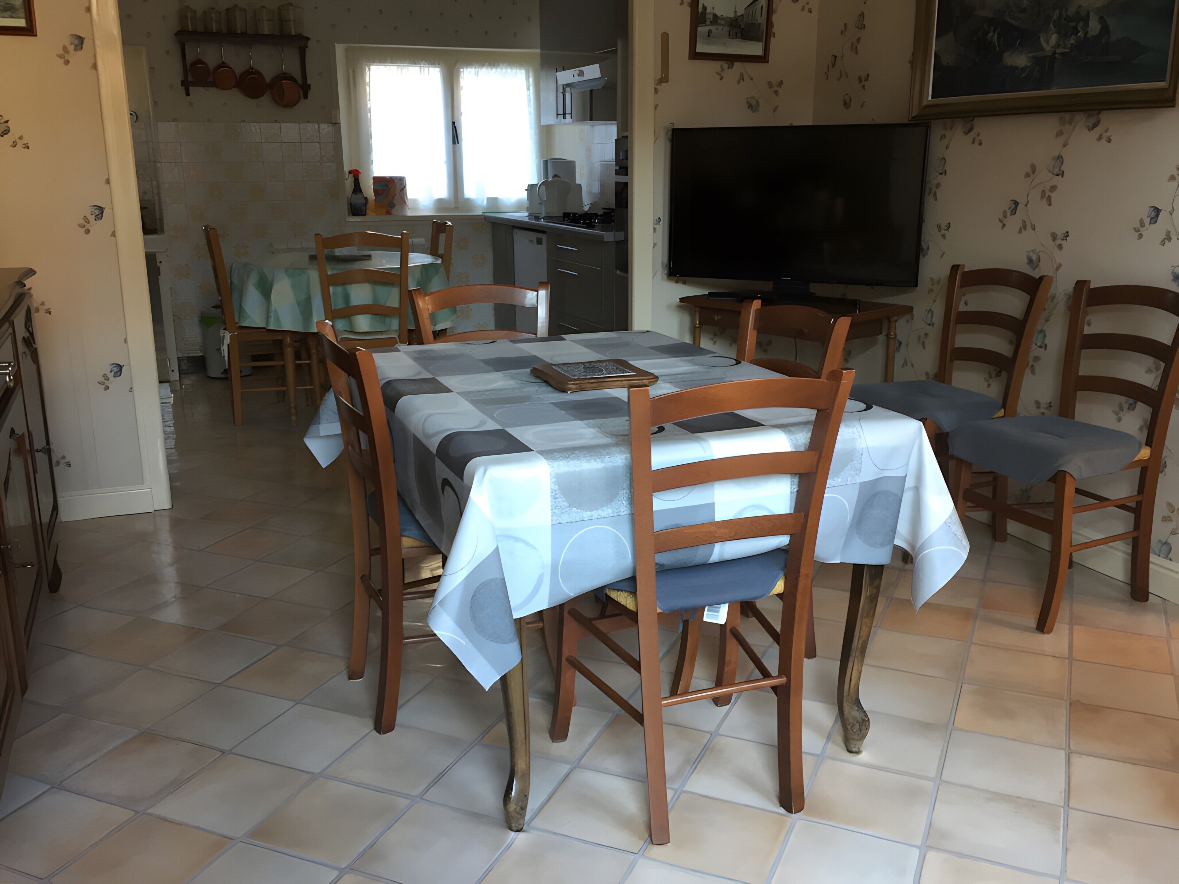 Uma sala de jantar com mesa posta para quatro pessoas, cadeiras de madeira, uma televisão e cozinha ao fundo.