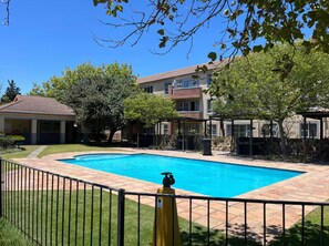 A swimming pool with a fenced area, surrounded by trees and a building with a tiled roof. - 2 Bedroom Pinelands Medical Leisure Apartment (Cape Town)