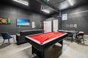 Une table de billard recouverte d’un drap de feutre rouge, un jeu de fléchettes et un écran de télévision diffusant une scène de plage.